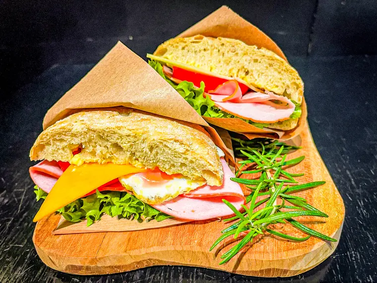 Breakfast ciabatta cut in half, filled with salad, cheese, ham and egg, served on a wooden board