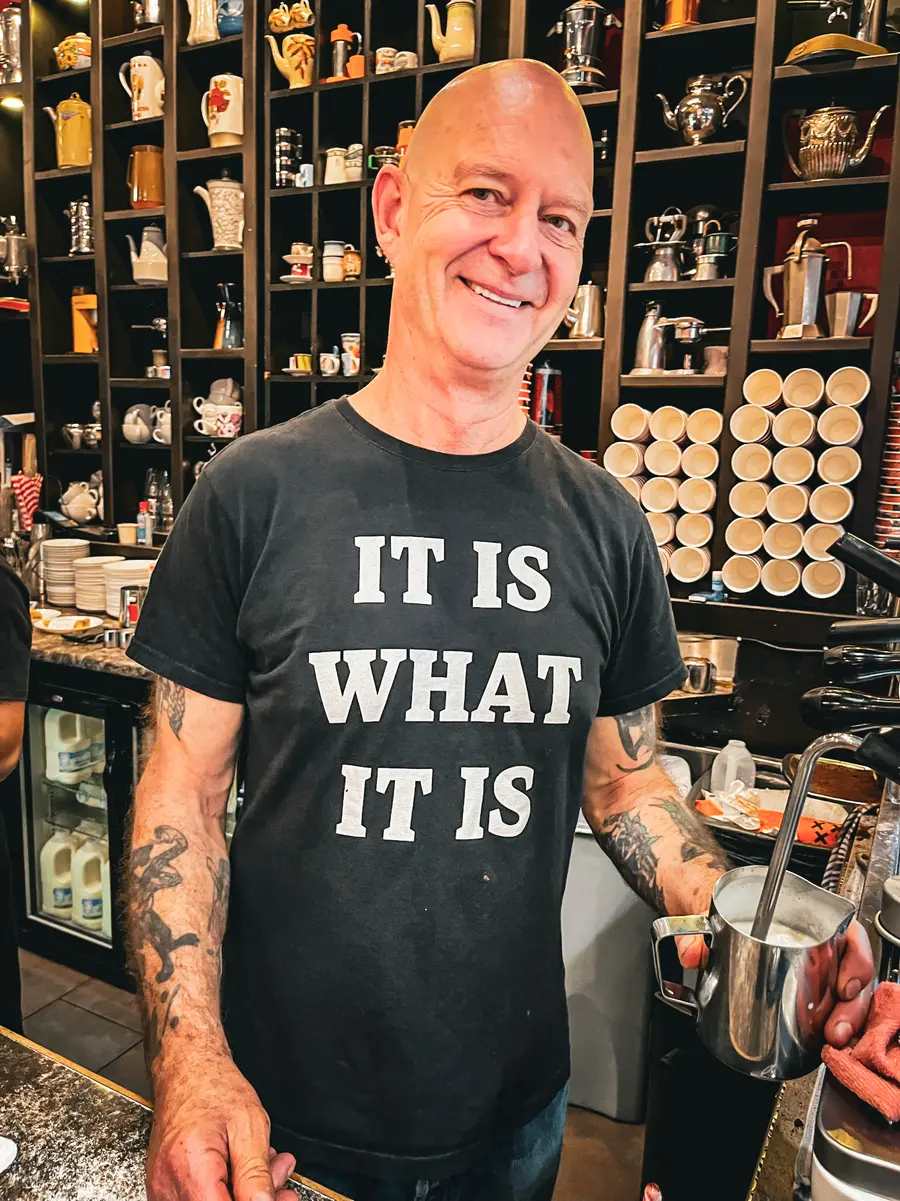 Buzz behind the espresso machine, wearing a t-shirt that reads It is what it is