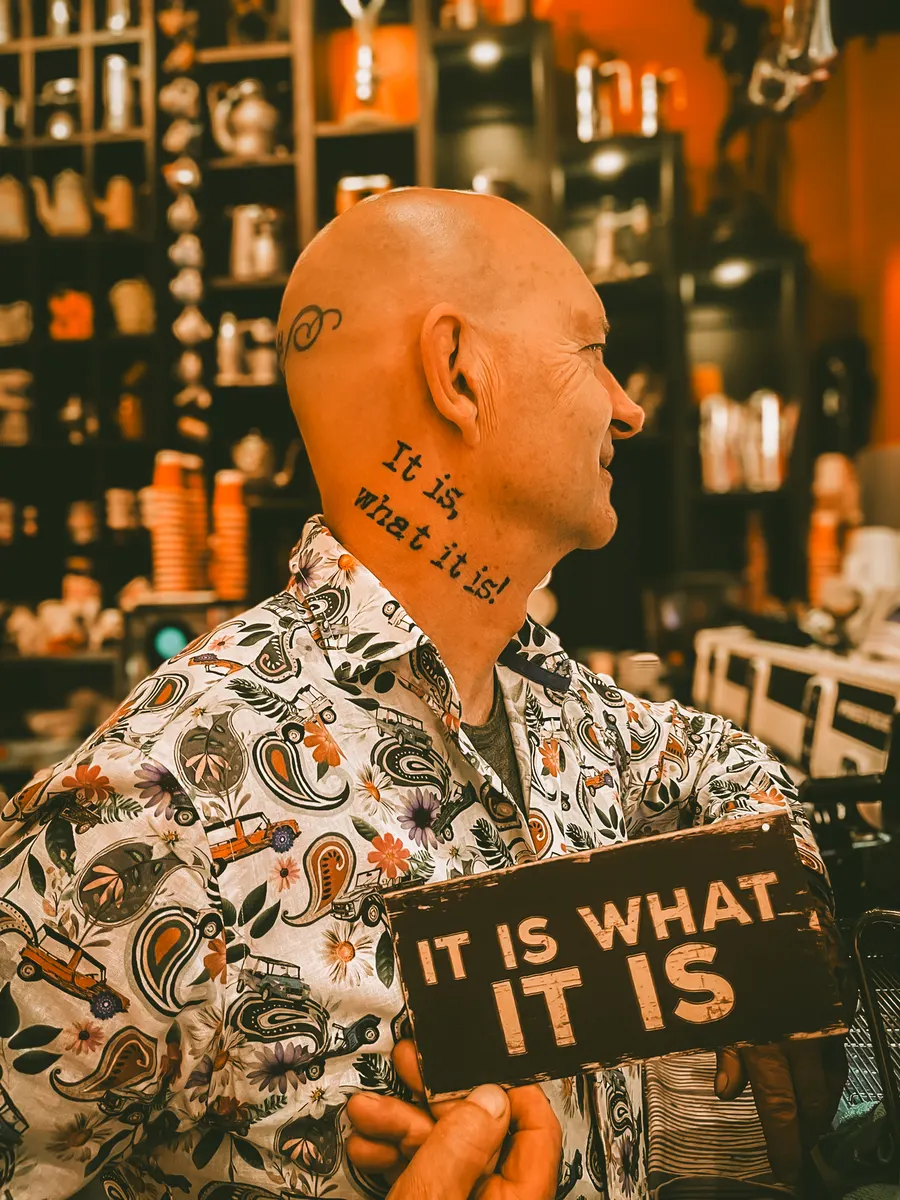 Buzz in profile in a floral shirt, the words It is what it is tattooed down the side of his head, holding a painted sign that says the same thing