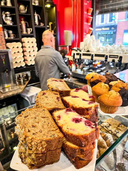 Slices of banana bread and raspberry sponge on plates at the counter, with muffins behind them