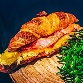 A croissant filled with scrambled egg and bacon, served on a wooden slice with rosemary