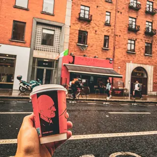 A takeaway cup held up across the street from the red Bald Barista shopfront on a wet day