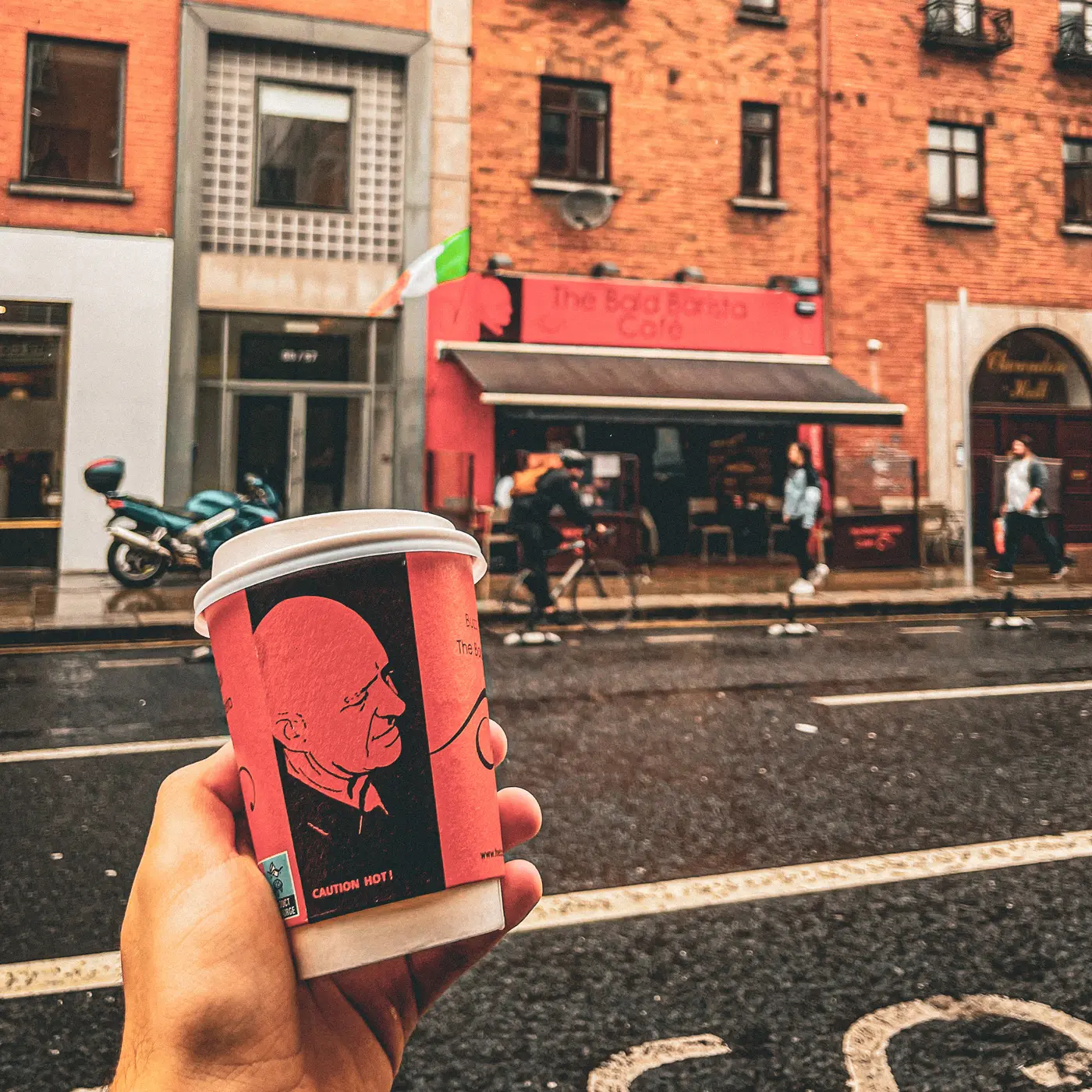A takeaway cup with Buzz's portrait held up in front of The Bald Barista shopfront on a wet morning on Aungier Street